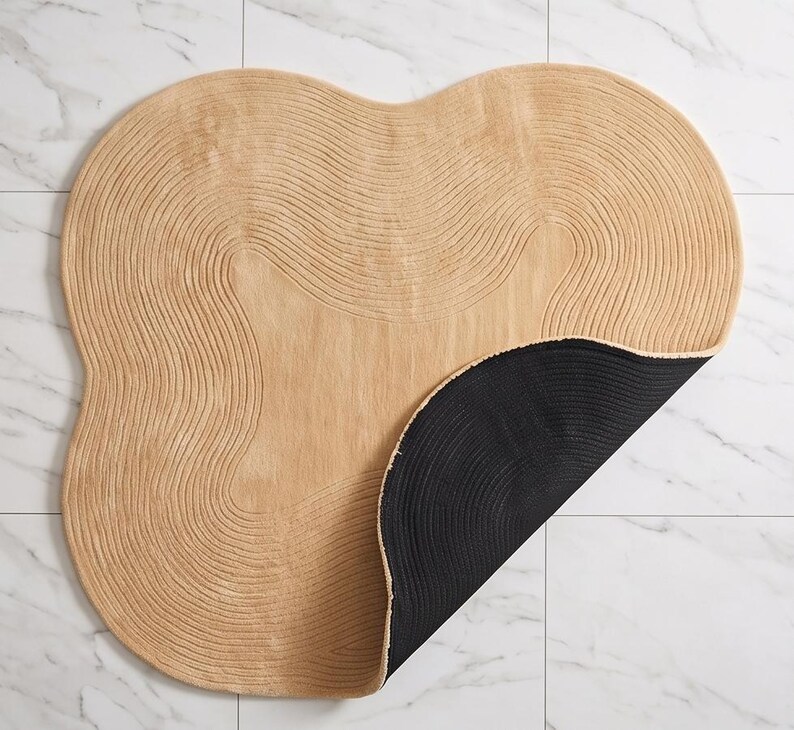 May include: An irregular, organic-shaped rug featuring a light tan, textured surface with concentric, wavy line patterns. The rug is partially folded over to reveal a solid black, circular-patterned underside, contrasting with the tan top.