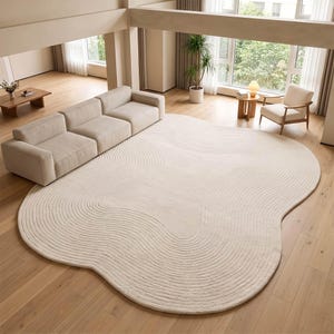 May include: An off-white area rug with an irregular, organic shape and a textured surface featuring concentric, wave-like carved lines. The rug is positioned on a light-colored wooden floor in a living room setting, partially underneath a modular sofa.