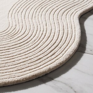 May include: An off-white area rug with a soft, textured surface featuring a series of raised, parallel, wavy lines that follow the curved, organic shape of the rug's edge, creating a rhythmic pattern across the entire fabric surface.