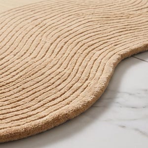 May include: A tan-colored rug with a curved, organic shape and a textured surface featuring raised, parallel wavy lines that create a rhythmic pattern across the entire piece, showcasing a soft, tufted pile construction throughout.