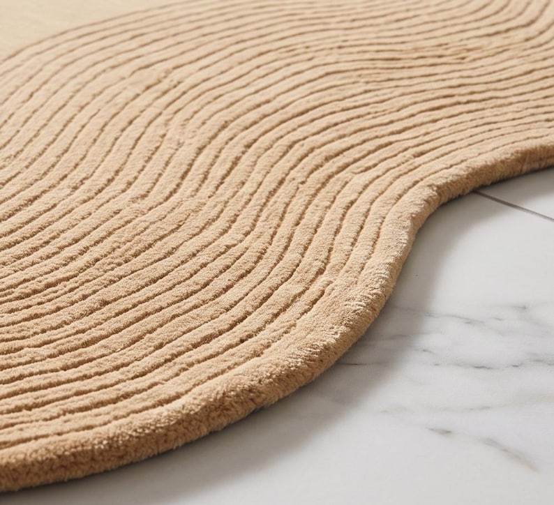 May include: A tan-colored rug with a curved, organic shape and a textured surface featuring raised, parallel wavy lines that create a rhythmic pattern across the entire piece, showcasing a soft, tufted pile construction throughout.