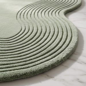 May include: A sage green area rug with an irregular, organic shape featuring a series of concentric, raised wavy lines that follow the curved perimeter of the piece, creating a textured, layered appearance across the entire surface.