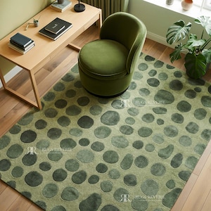 May include: A green area rug with a pattern of large, irregular circles in varying shades of green. A green velvet chair sits on the rug, near a wooden desk. The rug is on a hardwood floor.