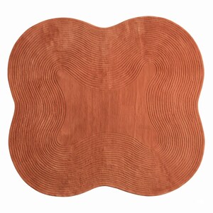 May include: A terracotta-colored area rug with a distinct four-lobed, organic shape. The surface features a series of concentric, raised wavy lines that follow the contour of the rug, creating a textured pattern across the entire piece.