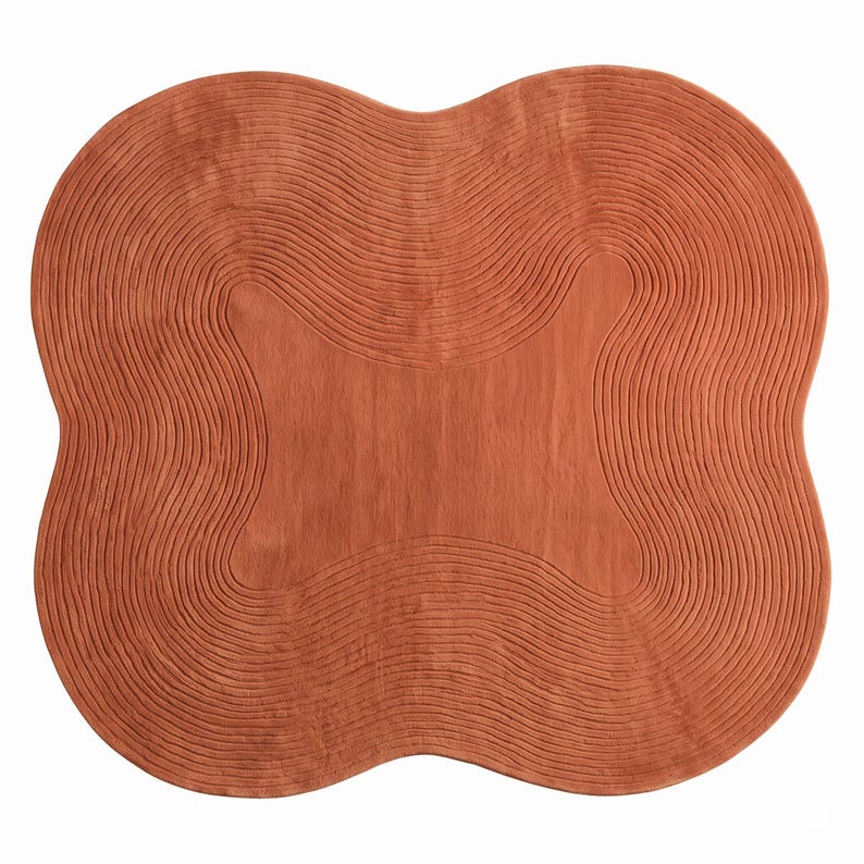 May include: A terracotta-colored area rug with a distinct four-lobed, organic shape. The surface features a series of concentric, raised wavy lines that follow the contour of the rug, creating a textured pattern across the entire piece.
