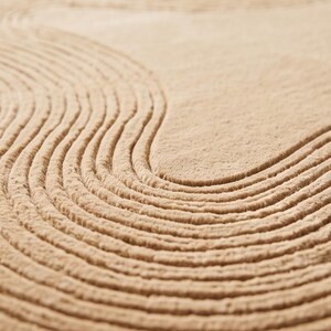 May include: A beige rug featuring a textured surface with carved, concentric wave patterns that create a series of raised, curved lines across the fabric. The material has a soft, dense pile with a uniform light tan color throughout the design.