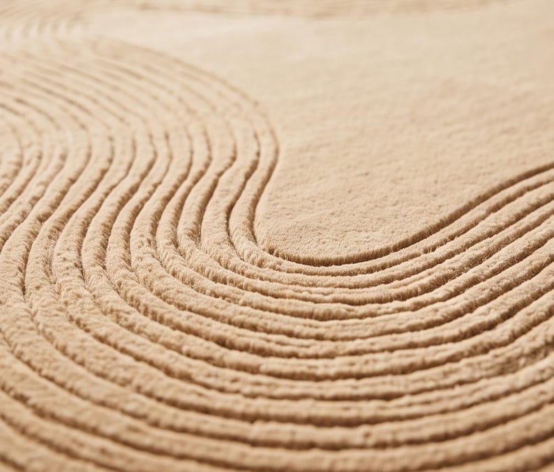May include: A beige rug featuring a textured surface with carved, concentric wave patterns that create a series of raised, curved lines across the fabric. The material has a soft, dense pile with a uniform light tan color throughout the design.