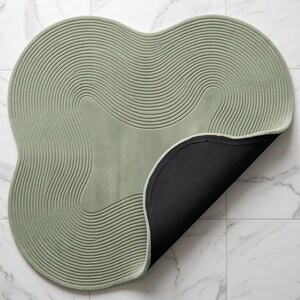 May include: A sage green rug with an irregular, organic shape and a textured surface featuring concentric, wavy line patterns. The rug is partially folded over to reveal a solid black, non-slip backing on the underside of the mat.