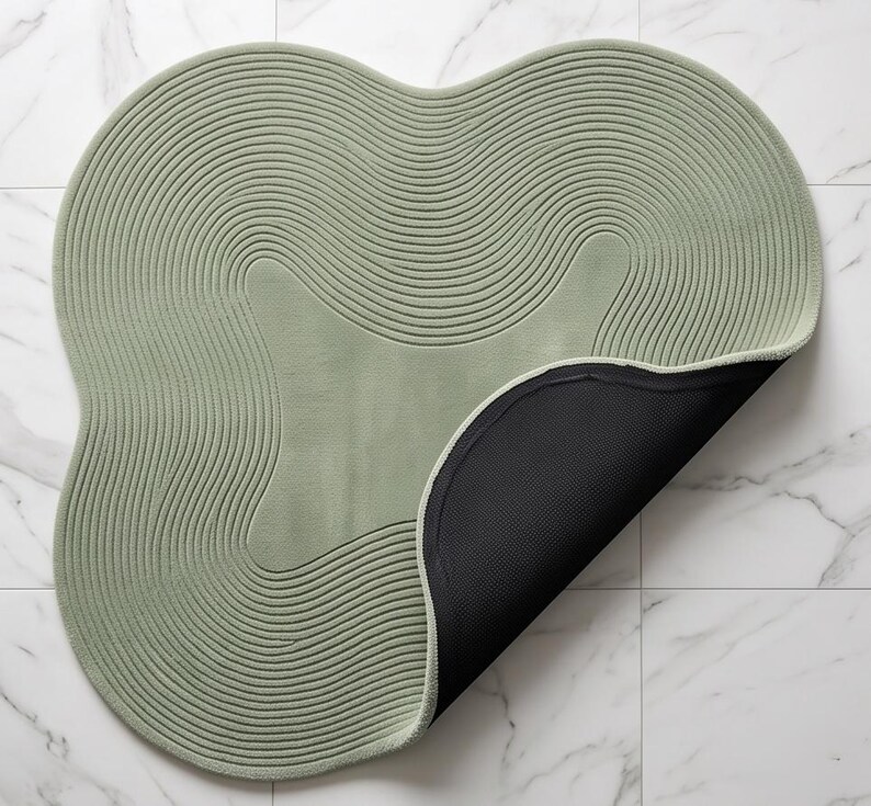 May include: A sage green rug with an irregular, organic shape and a textured surface featuring concentric, wavy line patterns. The rug is partially folded over to reveal a solid black, non-slip backing on the underside of the mat.
