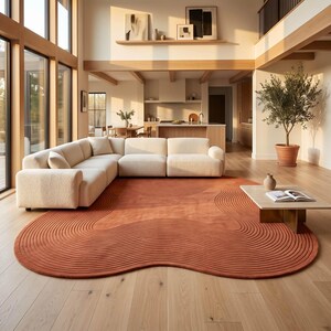 May include: A large, irregular, terracotta-colored area rug featuring a series of concentric, raised, wave-like lines that follow the organic shape of the rug, placed on a light-colored wooden floor in front of a cream-colored sectional sofa.