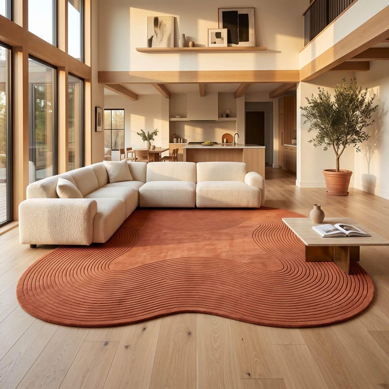 May include: A large, irregular, terracotta-colored area rug featuring a series of concentric, raised, wave-like lines that follow the organic shape of the rug, placed on a light-colored wooden floor in front of a cream-colored sectional sofa.