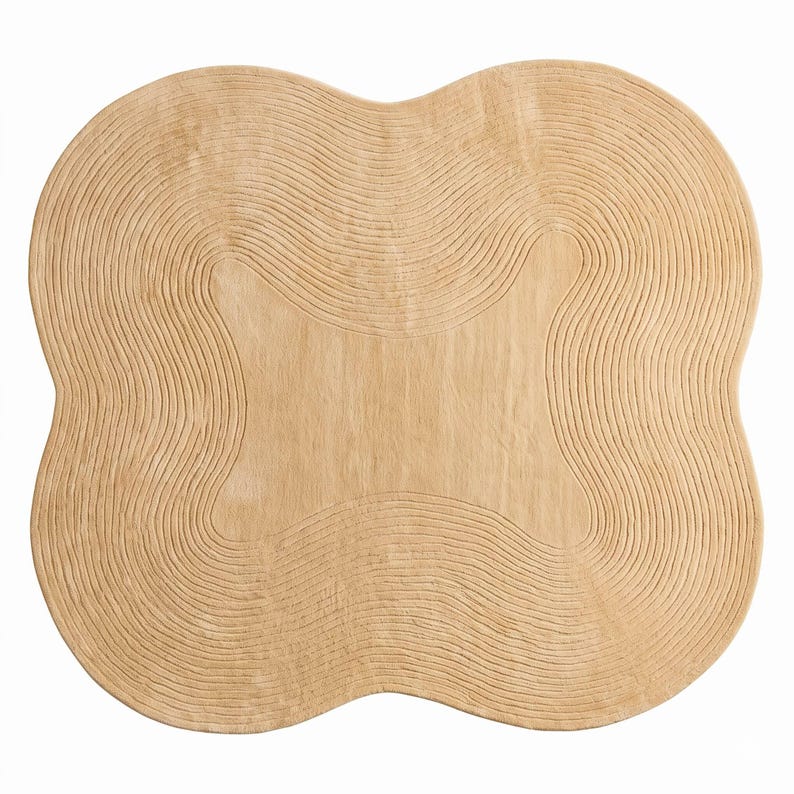 May include: A light beige rug with an irregular, organic four-lobed shape. The surface features a series of concentric, raised line patterns that follow the curved contours of the edges, creating a textured, rippled effect across the entire piece.