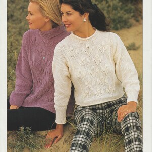 Può includere: Due donne con maglioni di lana. Uno è lilla chiaro, l'altro è bianco sporco con un motivo a foglie. Le donne sono sedute sulla sabbia. L'immagine contiene il testo "CHENILLE Double Knitting" e "SIRDAR".