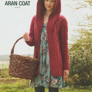 May include: A deep red hooded Aran coat with a cable knit pattern. The image shows a woman wearing the coat over a floral dress, holding a woven basket. The text on the image reads "Hooded Aran Coat (p. 24-27)".