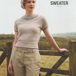 Könnte beinhalten: Eine Frau trägt einen Fair Isle Yoke-Pullover und eine karierte Hose. Der beige Kurzarmpullover hat kleine lila Punkte und einen floralen Ausschnitt. Die Hose ist hellbraun mit einem Karomuster. Der Text "FAIRISLE YOKE SWEATER" ist sichtbar.