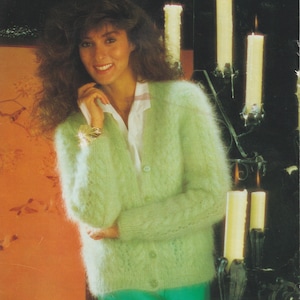 Könnte beinhalten: Eine Frau trägt einen hellgrünen, flauschigen Cardigan über einem weißen Hemd. Der Cardigan hat eine Knopfleiste und ein strukturiertes Muster. Der Hintergrund zeigt brennende Kerzen in dekorativen Haltern und eine orange-braune Wand.