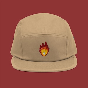May include: A tan five-panel cap with a flat brim. The cap features an embroidered flame design in red, orange, and yellow. The cap is made of a durable fabric and has a structured fit. The background is a solid maroon color.