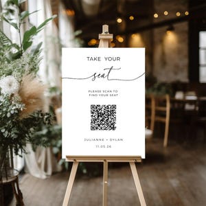 以下が含まれることがあります： 木製のイーゼルに立てられた白い看板。黒い筆記体で「TAKE YOUR seat」と書かれています。その下に「PLEASE SCAN TO FIND YOUR SEAT」と書かれ、QRコードが含まれています。「JULIANNE + DYLAN」という名前と日付「11.05.26」が一番下にあります。