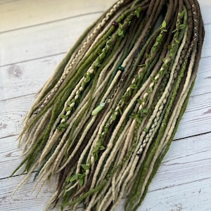 Puede incluir: Conjunto de rastas hechas a mano en tonos marrón, verde y crema. Las rastas están adornadas con vegetación artificial y costuras decorativas. La estética general es terrosa y bohemia, adecuada para peinados alternativos.