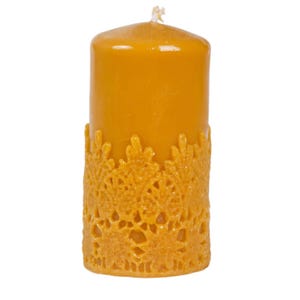May include: A cylindrical golden yellow pillar candle featuring a textured, raised lace-like pattern wrapped around the lower half. The candle has a smooth top surface with a single centered cotton wick extending from the middle of the wax.