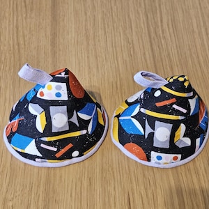 May include: Two cone-shaped fabric tea cosies featuring a black background with a colourful geometric pattern of circles, lines, and shapes in blue, yellow, orange, and white. Each cosy has a small light purple fabric loop at the top point.