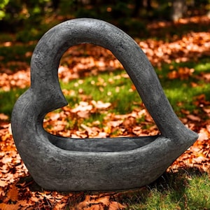 May include: A gray, heart-shaped planter made of stone. The planter has a textured surface and sits on a bed of fallen leaves and grass. The heart shape is open in the center, creating a unique design.