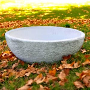 May include: A large, round, light gray stone planter bowl with a textured exterior. The interior is white. The planter sits on a bed of green grass and fallen autumn leaves, with a blurred background of grass and sunlight.