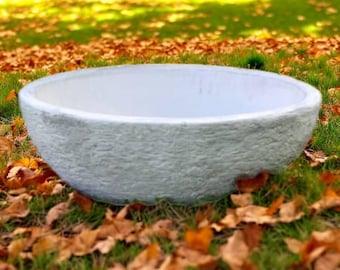 May include: A large, round, light gray stone planter bowl with a textured exterior. The interior is white. The planter sits on a bed of green grass and fallen autumn leaves, with a blurred background of grass and sunlight.