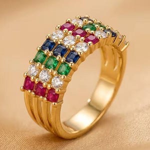 May include: A gold band ring featuring four parallel rows of square-cut gemstones. The rows consist of red, clear, green, and blue stones set in a multi-strand gold metal frame. The design showcases a vibrant, multi-coloured arrangement of gems.