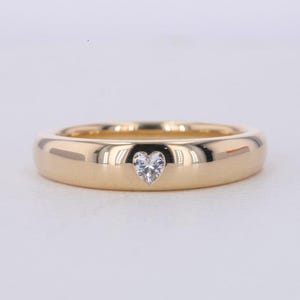 May include: A gold-colored ring with a heart-shaped diamond at the center. The ring has a smooth, polished surface and a rounded band. The diamond is clear and sparkling, set in a bezel setting. The ring is likely a piece of jewelry.