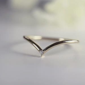 Op de afbeelding: Een delicate goudkleurige ring met een V-vormig ontwerp. De ring heeft een kleine, heldere edelsteen op het punt van de V. De ring staat tegen een zachte, vage achtergrond, wat de elegante eenvoud benadrukt.