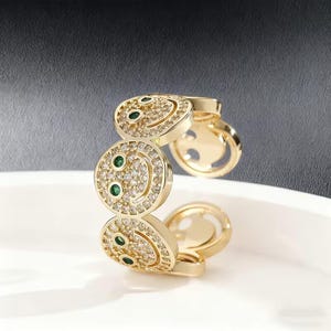 Puede incluir: Un anillo de tono dorado que presenta una serie de motivos circulares de caras sonrientes conectadas. Cada cara está incrustada con pequeñas piedras claras e incluye dos pequeñas piedras redondas de color verde vibrante como ojos.