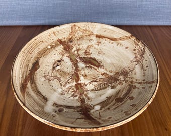 以下が含まれることがあります： ベージュのベースと茶色の抽象的なスプラッターデザインが施された、丸いセラミック製のサービングボウル。ボウルにはわずかに盛り上がった縁があり、木製の表面に置かれています。ボウルの内側には同心円があります。