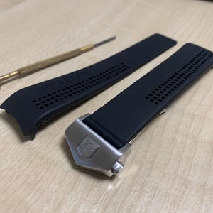 May include: Black watch strap with a silver clasp, a small screwdriver, and a second strap piece. The watch strap has a perforated design and the brand name "TAG HEUER" is visible on the strap.