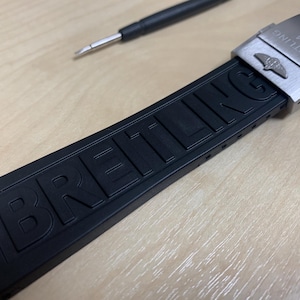 May include: Black rubber watch strap with the brand name "BREITLING" embossed on the band. The strap has a stainless steel buckle with the Breitling logo and the text "BREITLING 1884". A small black tool is in the background.