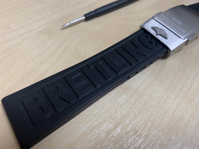 May include: Black rubber watch strap with the brand name "BREITLING" embossed on the band. The strap has a stainless steel buckle with the Breitling logo and the text "BREITLING 1884". A small black tool is in the background.