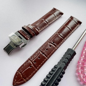 May include: Brown leather watch straps with a crocodile pattern, a silver clasp with the Longines logo, and a black tool. The straps are new and unused. A pink beaded bracelet is also in the image.