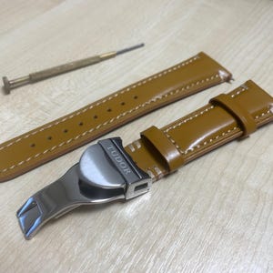 Puede incluir: Una correa de reloj y un juego de herramientas. La correa del reloj es de cuero marrón claro con costuras blancas. El cierre es plateado y tiene la palabra "TUDOR" grabada. También se incluye un pequeño destornillador.
