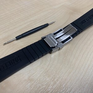 May include: A black rubber watch strap with a stainless steel clasp and a small black screwdriver. The strap has the word "BREITLING" embossed on it. The watch strap is on a light brown wooden surface.