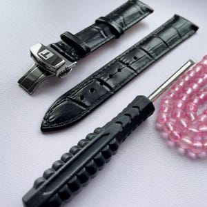 Peut inclure: Bracelet de montre en cuir noir avec fermoir argenté, un outil noir pour bracelet de montre et un brin de perles roses. Le bracelet de montre a un motif peau de crocodile.