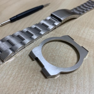 May include: A collection of watch components, including a stainless steel watch strap, a watch case, and a tool, are displayed on a light wooden surface. The watch strap is made of brushed metal links. The case is a circular frame.
