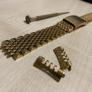 May include: A gold-colored watch band with a mesh design, along with two detached end pieces and a small screwdriver. The watch band is made of interlocking metal links. The items are on a light-colored surface.