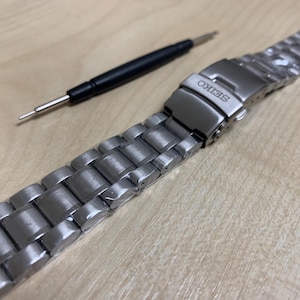 May include: A stainless steel watch band with a deployment clasp, featuring the brand name "SEIKO". A watch band adjustment tool is also present. The band is made of interlocking metal links.