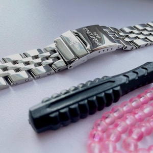 Könnte beinhalten: Ein Edelstahl-Uhrenarmband mit Faltschließe, auf der das Wort "BREITLING 1884" eingraviert ist. Ein schwarzer Schraubendreher und eine Schnur aus rosa Perlen sind ebenfalls im Bild. Das Armband besteht aus ineinandergreifenden Metallgliedern.