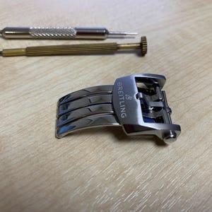 May include: A silver-colored watch clasp with the brand name "BREITLING" engraved on it. Two watch repair tools are in the background. The clasp has a curved design with multiple parallel ridges.