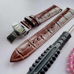 May include: Brown leather watch straps with a crocodile pattern, a silver clasp with the Tissot logo, a black tool, and a strand of pink beads. The watch straps are new and ready to be installed.