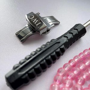 May include: A stainless steel watch clasp with the IWC logo, a black screwdriver, and a strand of pink beads. The clasp is open, showing its inner workings. The screwdriver has a textured handle. The beads are a light pink color.