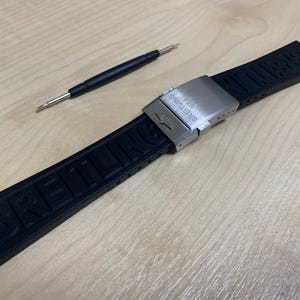 May include: A black rubber watch strap with the brand name "Breitling" embossed on it. The strap has a silver-colored clasp and a small tool for adjustment. The items are on a light-colored wooden surface.