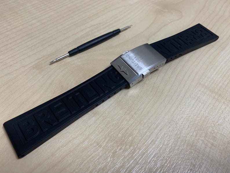 May include: A black rubber watch strap with the brand name "Breitling" embossed on it. The strap has a silver-colored clasp and a small tool for adjustment. The items are on a light-colored wooden surface.