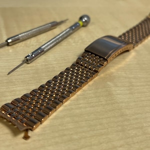 May include: A rose gold-toned metal watch band featuring a multi-link design with a folding clasp, accompanied by a silver-toned metal spring bar tool with a textured grip, used for adjusting or removing watch pins from the bracelet links.